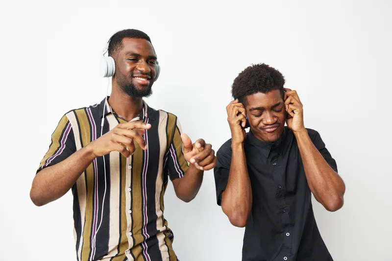 Two happy black men enjoying music together, wearing headphones and smiling with joy. Their vibrant outfits reflect a casual and fun atmosphere.