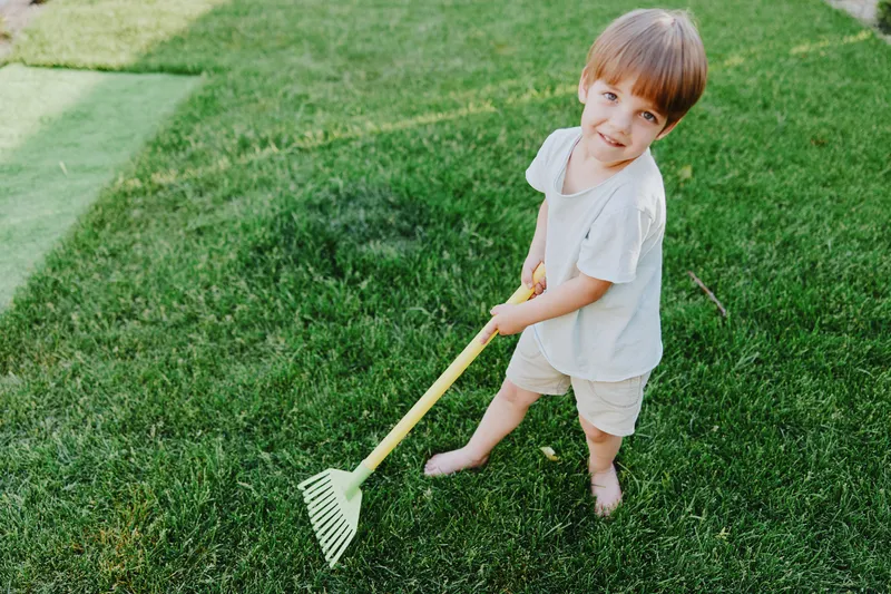 Playful boy working in the garden, enjoying outdoor activities with a bright smile, wearing casual clothes and standing on lush green grass in the sunlight.