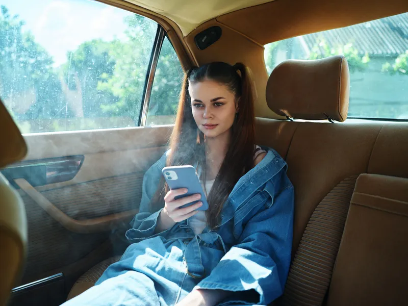 Young woman in denim outfit, sitting in the backseat of a car, using her smartphone, with a relaxed expression and natural sunlight creating a warm ambiance.