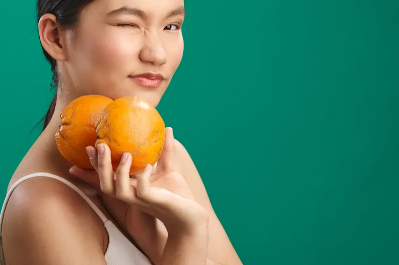 asian woman holds oranges near her face against a green backdrop, a gentle smile and calm expression create a fresh, relaxed portrait with a healthful lifestyle mood.
