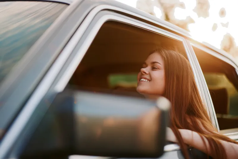 Happy young woman enjoying a summer drive with the window down, sunlight illuminating her face and long hair in warm, natural tones.