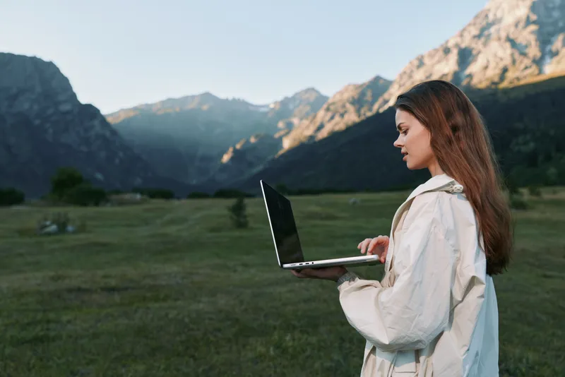 A focused professional woman uses a laptop outdoors in a scenic mountain landscape, conveying remote work, concentration, and modern productivity amid nature.