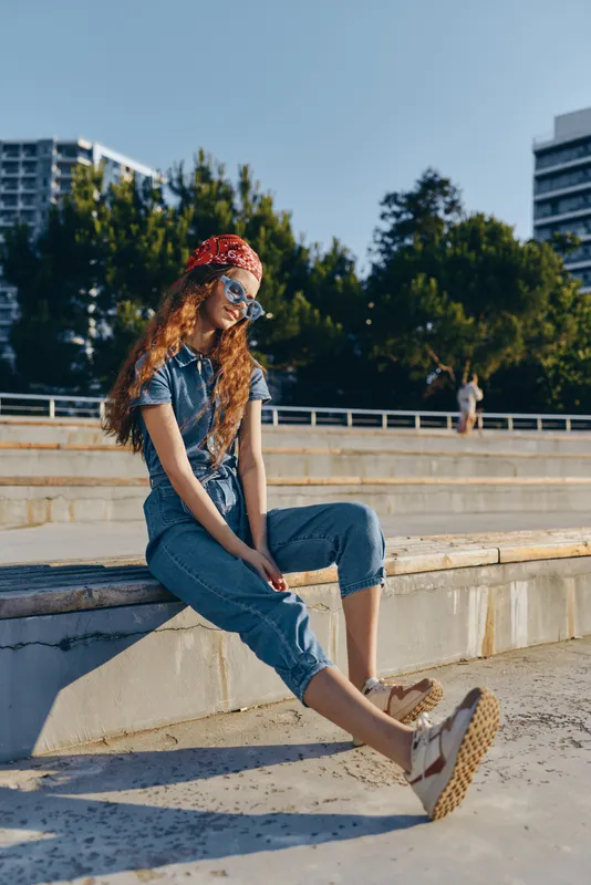 Lifestyle woman in modern boho-western denim street style with red bandana and sunglasses, casual relaxed pose outdoors in urban setting with film color effect