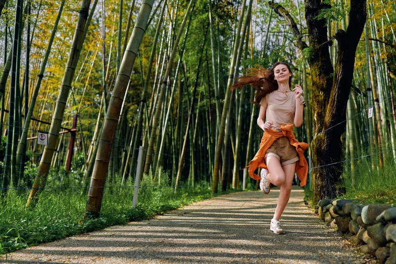 Woman running on a bamboo path in forest, joyful young woman jogging along a sunlit trail, casual summer outfit, flowing hair and carefree energy among tall green bamboo scenic.