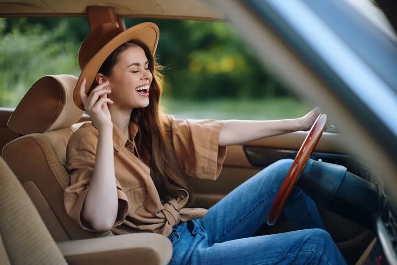 Young woman enjoying a road trip, wearing a stylish hat, smiling in a vintage car with a blurred green background.