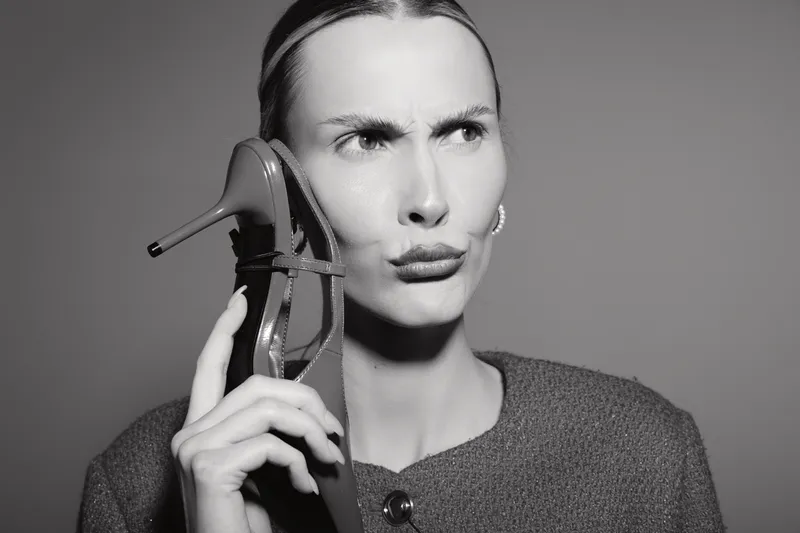 A fair skinned Caucasian woman with blonde hair holds a high heel shoe to her ear, looking confused and thoughtful against a plain background. Concept of creativity and humor.