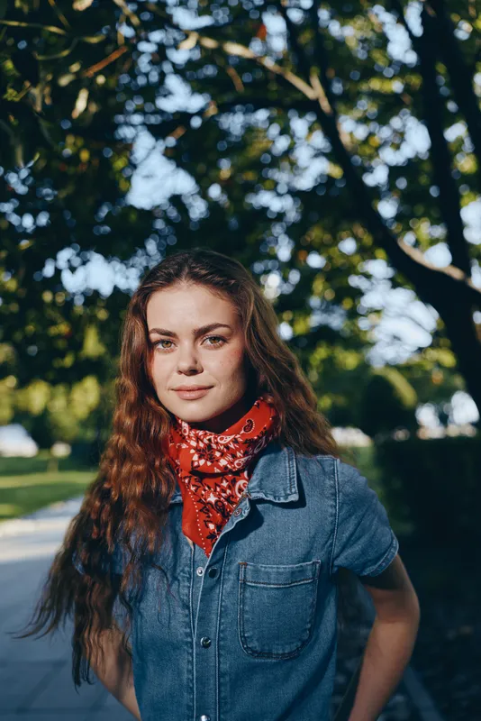 lifestyle woman in modern boho-western denim street style wearing red bandana with natural makeup standing outdoors under bright sunlight in film color