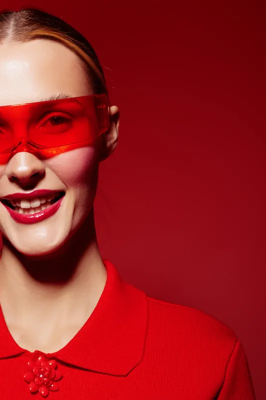 Smiling woman with red sunglasses and red shirt poses against red background. Close-up portrait with bright makeup and confident expression. Concept of fashion, style, and color contrast.