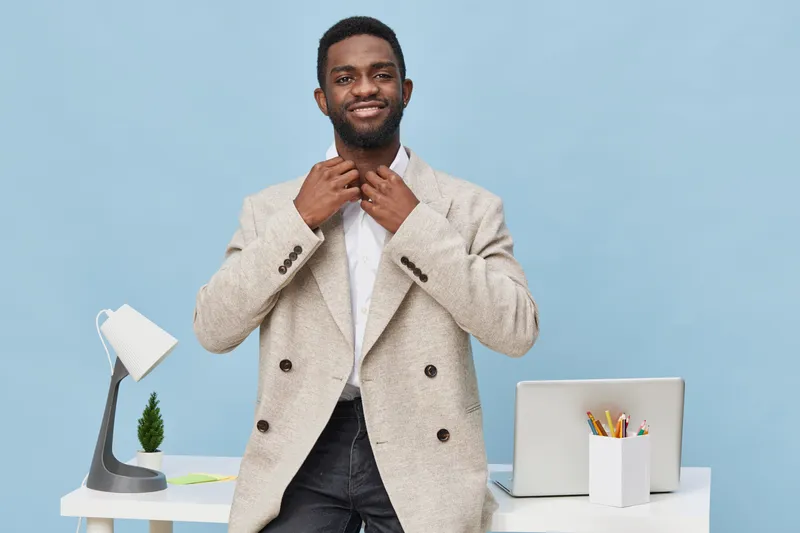 Confident smiling man with dark skin, casual beige blazer, white shirt, standing in modern office with light blue background, relaxed posture, professional appearance, interior background, studio shot