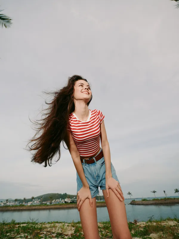 Young woman with long flowing hair wearing a red striped tee and denim shorts leans forward with a joyful smile, sunlit outdoor setting by water and open sky.