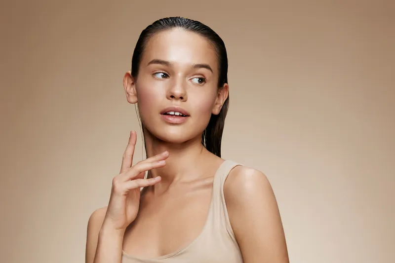 Young female model with wet hair in a neutral tank top, showcasing a natural and serene expression against a soft beige background, ideal for beauty and skincare concepts