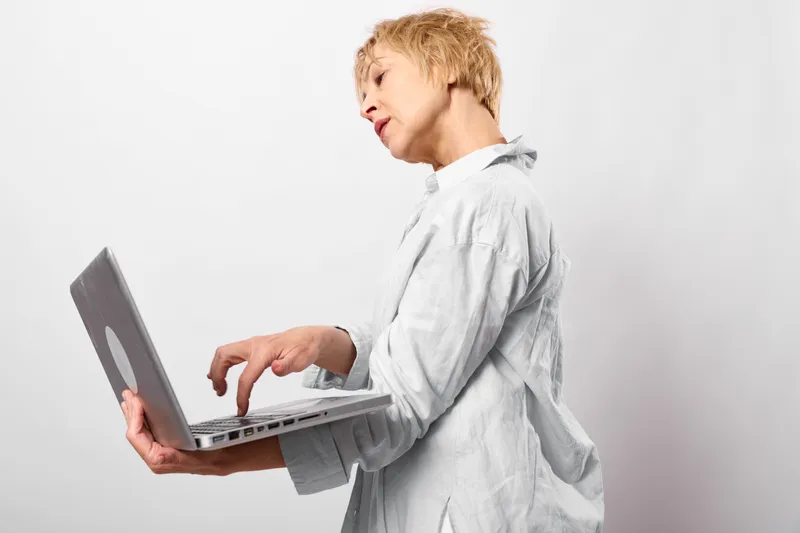 Stressed woman in white coat holds a laptop, hand on neck. Healthcare burnout and anxiety for mental health pressure in a calm studio with pure bright light.