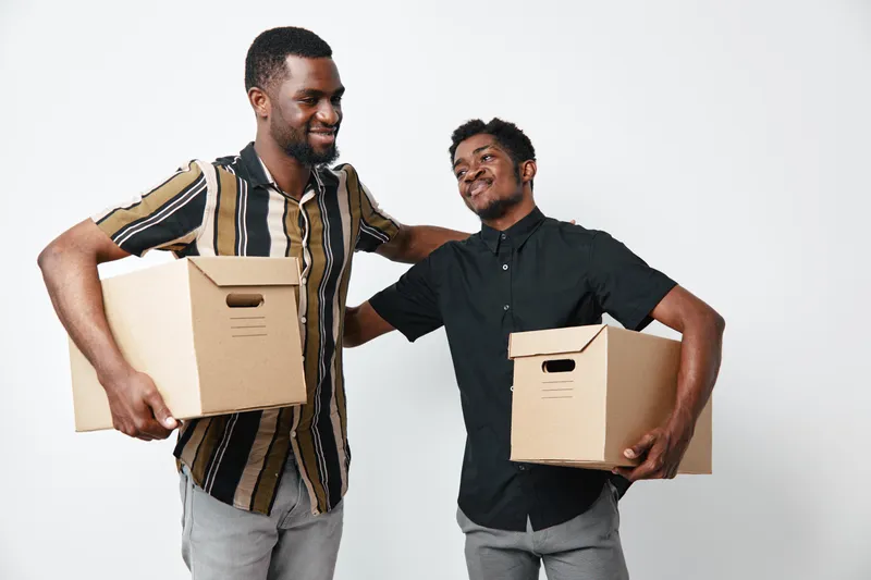 Two African American men with dark skin holding cardboard boxes smiling and interacting on white background for moving or delivery concept.