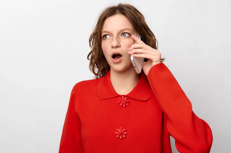 Surprised woman in red sweater talks on smartphone with open mouth, looking aside. Isolated on white background. Concept of unexpected news, communication, and emotions.