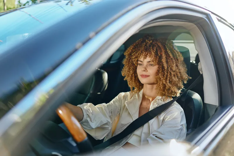 Smiling woman driving a modern car, wearing casual light-colored clothing and seatbelt, enjoying daylight and relaxed mood with curly hair and natural makeup inside the vehicle.