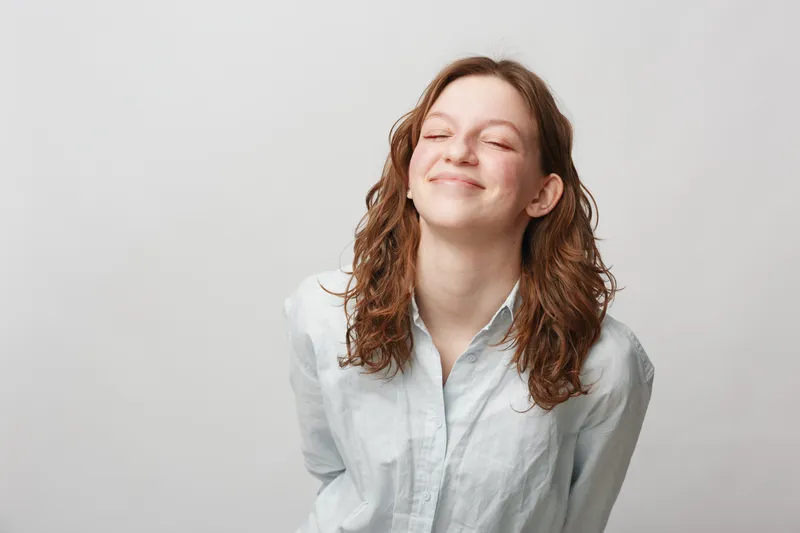 Young woman with curly hair smiles with closed eyes wearing white shirt on light background. Concept of happiness, relaxation, and positive emotions in casual lifestyle.