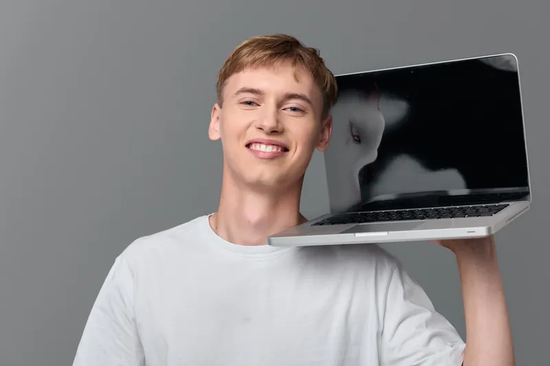Young man balances a laptop on his shoulder with a bright smile, casual tee, modern tech vibe, and a relaxed workspace feel that highlights everyday computing and digital presence for contemporary