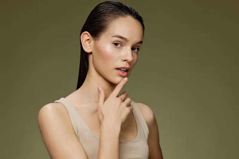 Portrait of a thoughtful young woman with straight hair on a green background, wearing a beige top and expressing curiosity and confidence