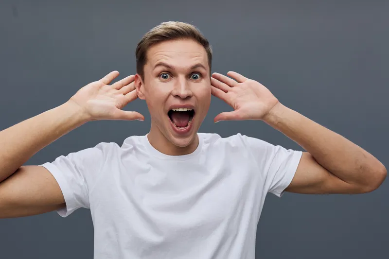 Excited young caucasian man with open mouth white t-shirt raised hands dark gray background expression emotion concept mental health gadgets