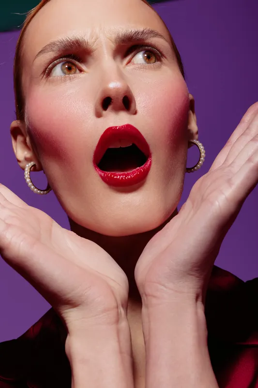 Surprised woman with red lipstick and hoop earrings looks shocked with open mouth and raised hands on purple background. Concept of emotion, beauty, and facial expression.