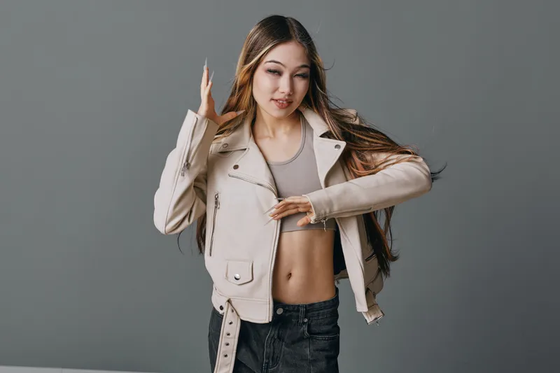 Young woman with long hair wearing beige jacket and crop top posing with hand near face isolated on grey background casual fashion style and confident look studio portrait.
