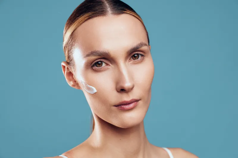 Closeup portrait of young woman with clear skin applying moisturizing cream on cheek looking at camera with neutral expression isolated on blue background.
