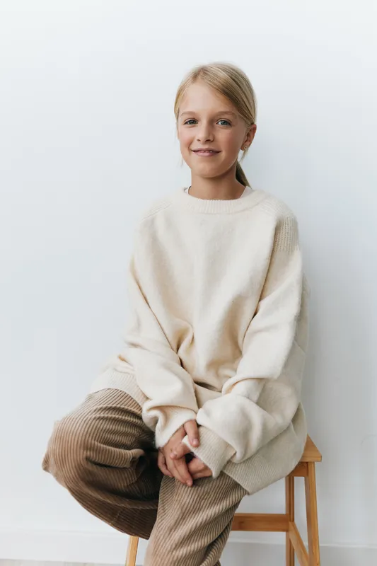 Portrait of a young girl seated on a wooden stool, wearing a soft beige knit sweater and corduroy pants, radiating calm and quiet confidence in a minimalist studio.