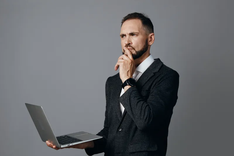 A professional man in a tailored suit stands against a solid colored background, holding a laptop and wearing a thoughtful, focused expression that conveys curiosity and leadership.