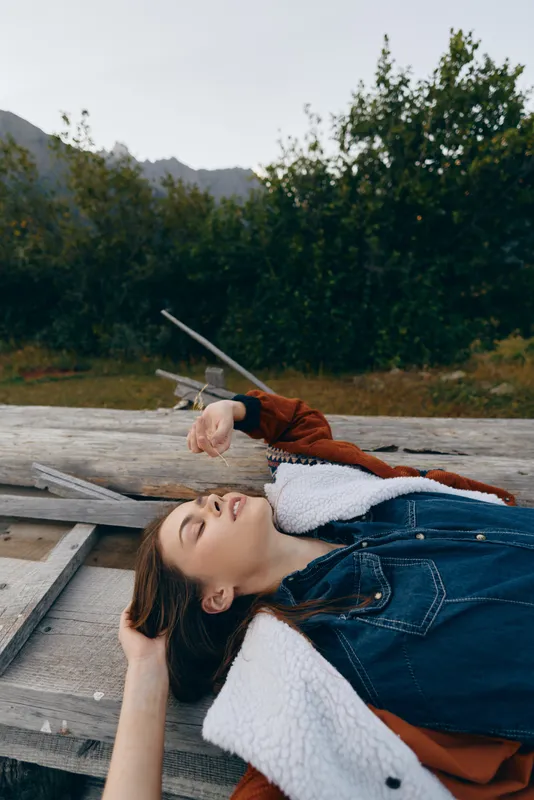 Woman lying outdoors on wooden plank, portrait of relaxation and calm in nature. Casual denim and shearling coat, peaceful expression, autumn landscape and soft light mood.
