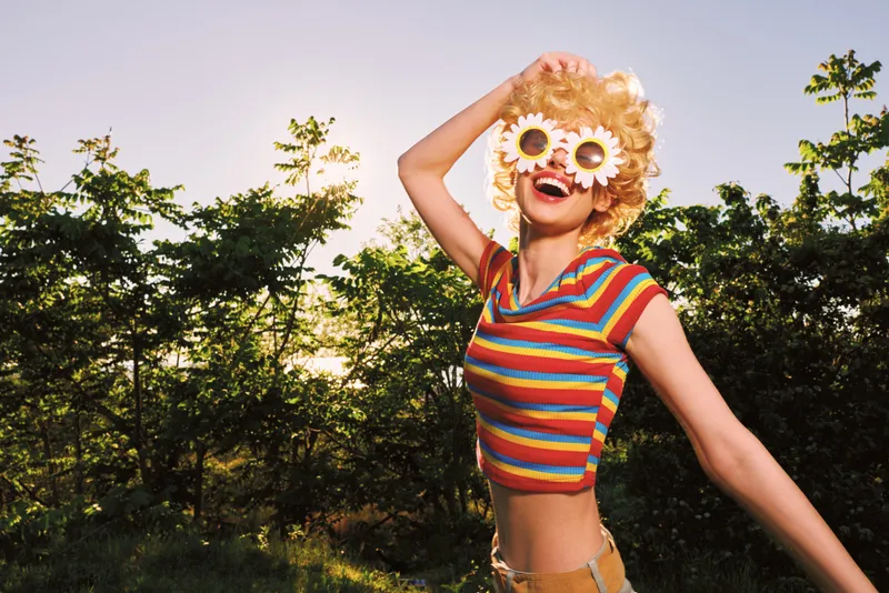Smiling woman outdoors wearing a striped crop top and flower sunglasses in a sunny field with retro film color tones. Casual lifestyle scene, carefree mood, warm light.