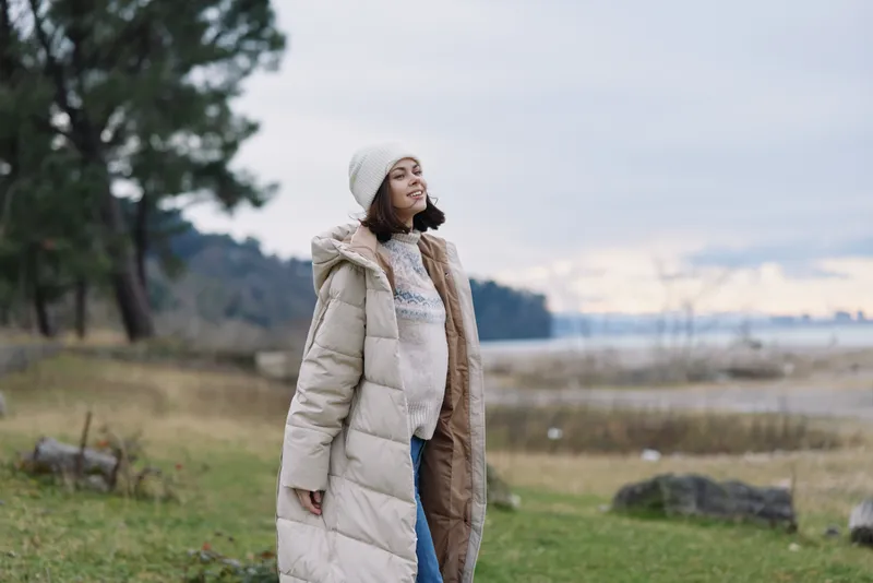 Woman in a long puffer coat standing in a lakeside park, chilly weather, trees in the background, calm outdoor moment with soft natural light.