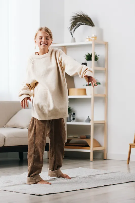 A cheerful child dances in a cozy sweater and soft pants, barefoot on a light rug, capturing joyful movement and home comfort.