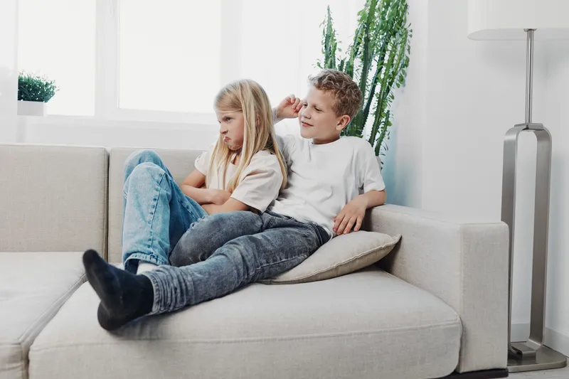 Two children sit on a couch, a boy playfully teasing a girl who looks upset, in a bright, modern living room setting.