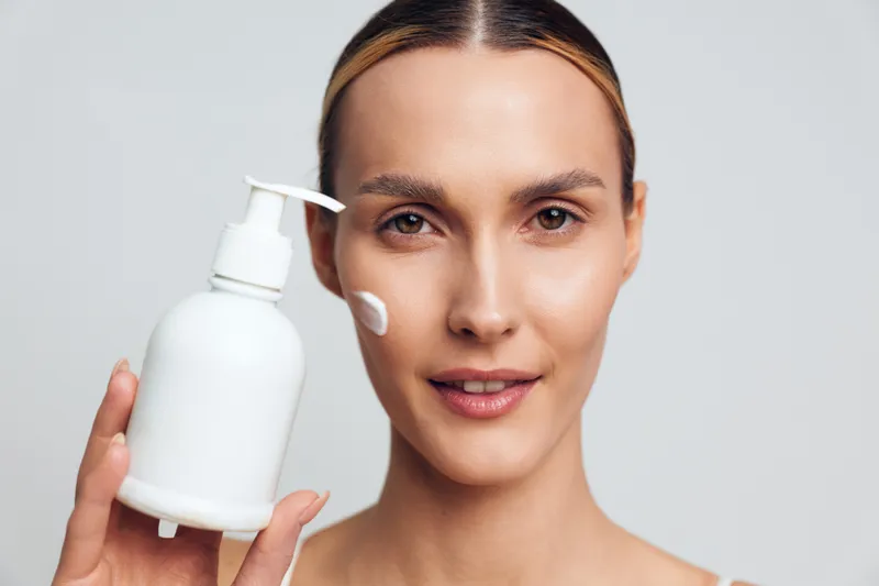 Young woman with clean skin applies moisturizing cream on face holding white plastic pump bottle isolated on gray background with natural beauty and skincare concept.