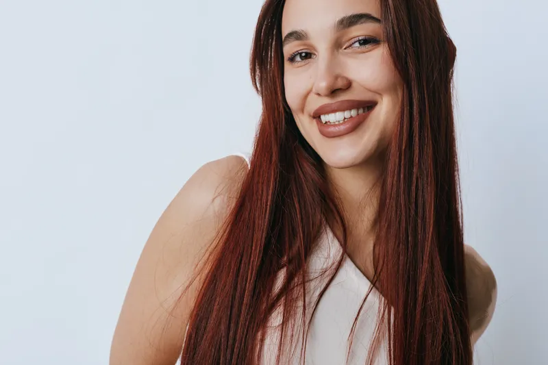 Young woman with long brown hair smiling and looking at camera in closeup portrait. Natural beauty, casual style and positive expression on white background with soft lighting.