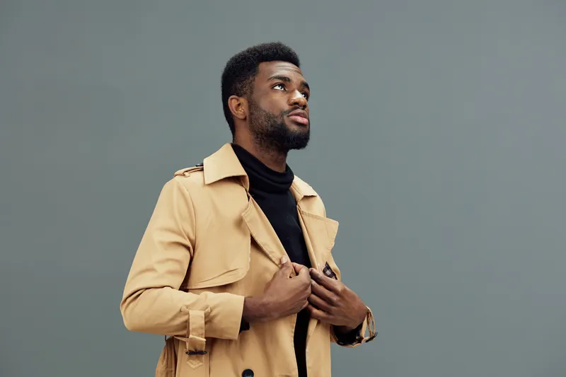 Stylish young man in a beige trench coat, looking up with a thoughtful expression against a grey background, embodying modern fashion and confidence, ideal for lifestyle concepts