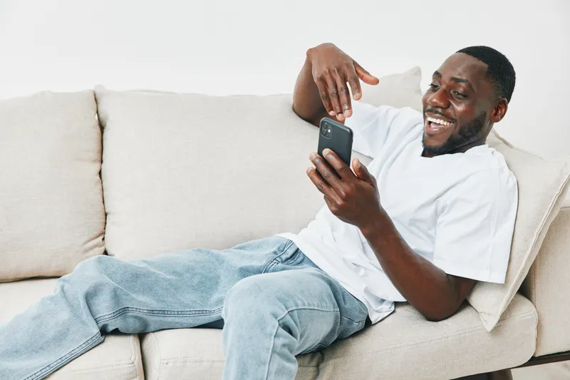 Young African man smiling while using a smartphone on a cozy couch in a bright living room, showcasing relaxation and joy in casual attire