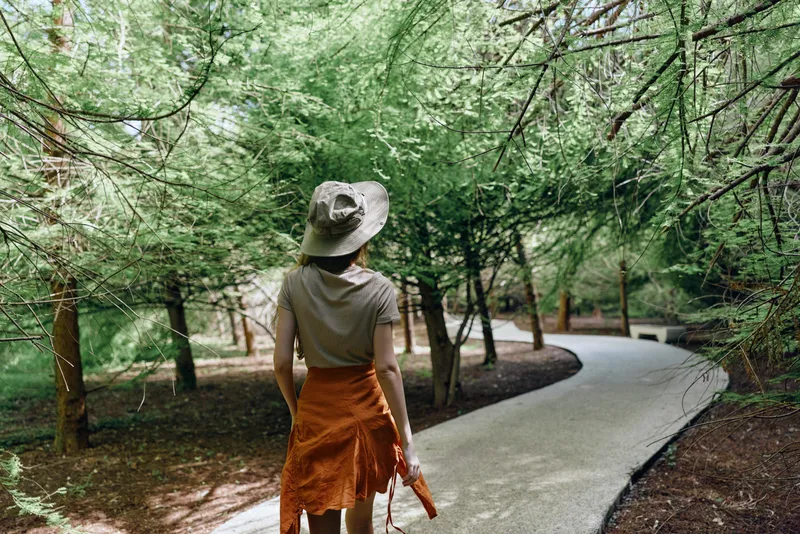 Woman walking on a winding path through a sunlit forest wearing a wide hat and orange skirt, backview portrait of casual outdoor leisure with trees, foliage and dappled sunlight on trail.