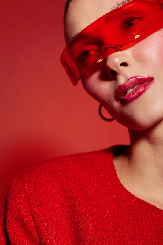 Close-up of a woman wearing red transparent glasses and a red sweater against a red background. Concept of fashion, style, and modern accessories with bold color contrast.