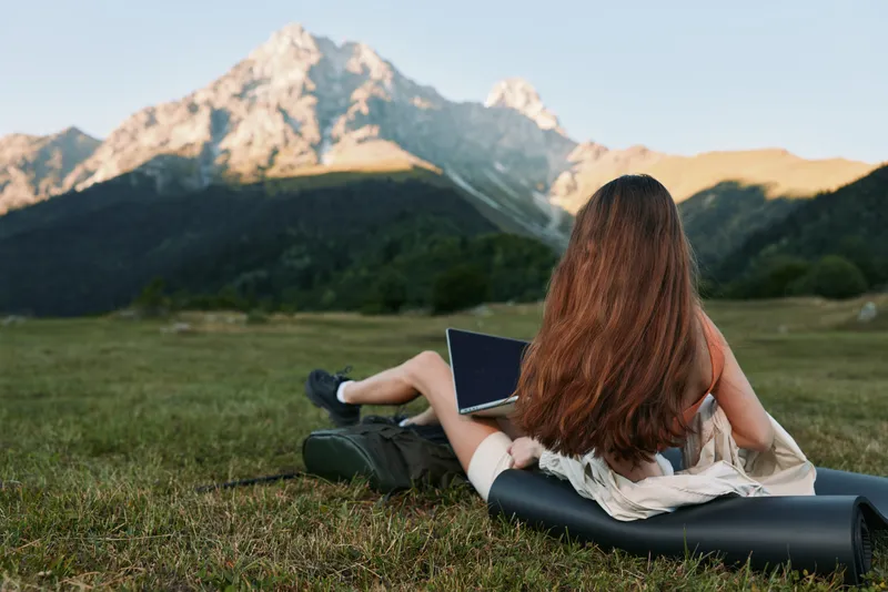 A person relaxes outdoors with a laptop, lying on a mat in a grassy meadow. Distant mountains frame a serene, productive work environment.