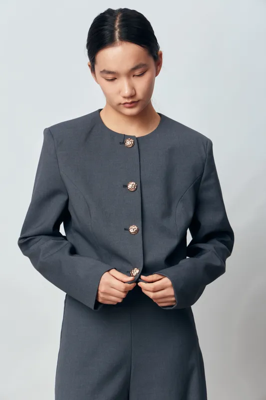 Young woman in gray tailored suit with decorative buttons buttoning jacket, standing indoors on plain background. Fashion portrait of professional confident businesswoman in elegant outfit.