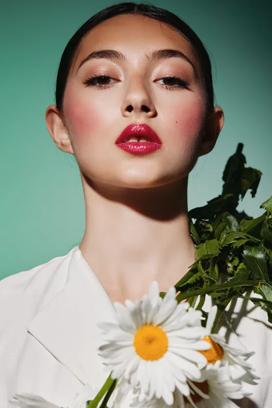 Close-up portrait of confident young woman with glossy red lipstick and natural finish skin holding white daisies on colored background. Creative concept of beauty fashion makeup.