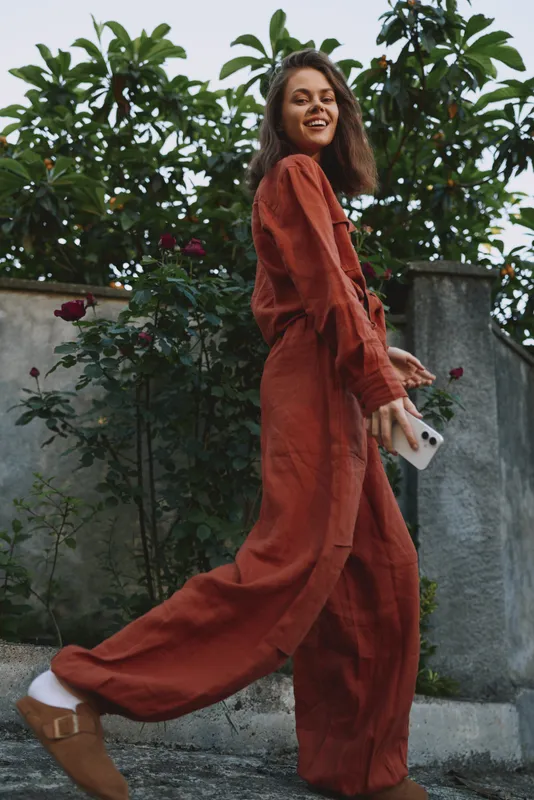 Young woman in a rust jumpsuit walks outdoors, smiling, holding a phone, against a stone wall and greenery, creating a lively casual lifestyle moment