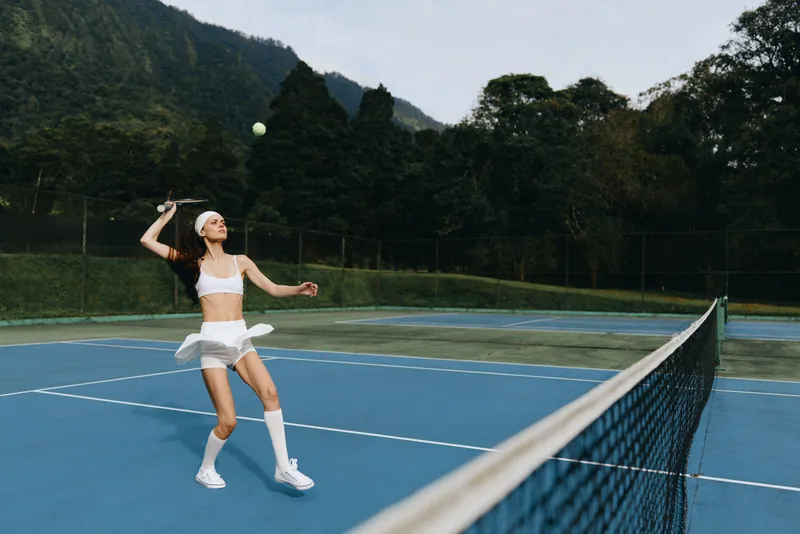 Young female tennis player in white sportswear, playing on a blue tennis court surrounded by greenery, embodying energy and enthusiasm for a healthy lifestyle