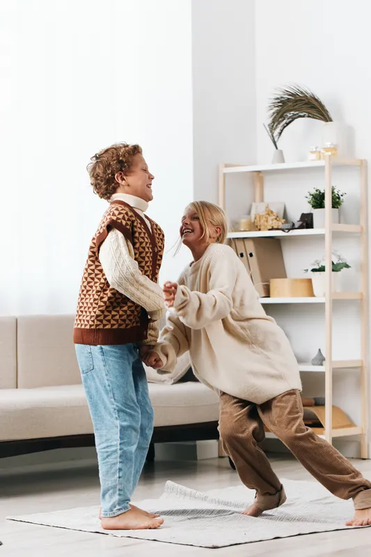 Two children laugh and chase each other indoors, a playful moment captured barefoot on a light rug in a cozy living room, radiating pure happiness.