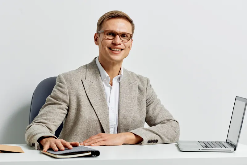 Smiling man wearing stylish glasses and a beige blazer sitting at a desk with laptop and notebook in a modern office environment, conveying professionalism, confidence and success in a business settin