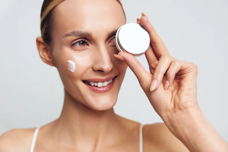 Young woman with clean skin applies face cream on cheek holds jar of moisturizer smiles gently isolated on white background beauty skincare concept.