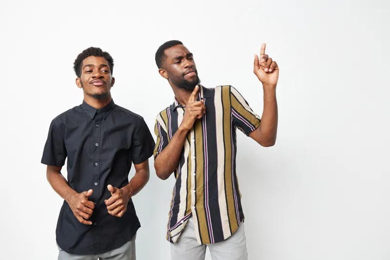 Two smiling Black men pose playfully against a light background, showcasing friendship and joy. Their vibrant outfits highlight their style and personality.