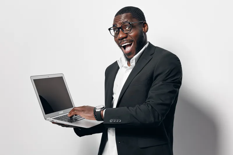 African American man with dark skin wearing glasses and black suit holding laptop with excitement on white background. Business professional, technology, communication and success concept.