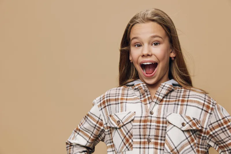 Happy girl with long hair wearing casual checkered shirt smiling and showing excitement with open mouth on beige background. Child with emotion and positive expression portrait.